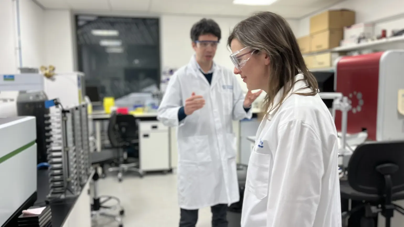 Angie, a Programme Director, in a lab coat and goggles in a lab during discovery. There is also a person in the background speaking and gesturing with their hands. 